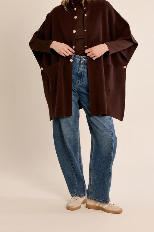 A woman modeling a dark brown open-front poncho-style cardigan with gold-tone buttons, worn over a black top and blue jeans, against a solid light beige studio background.