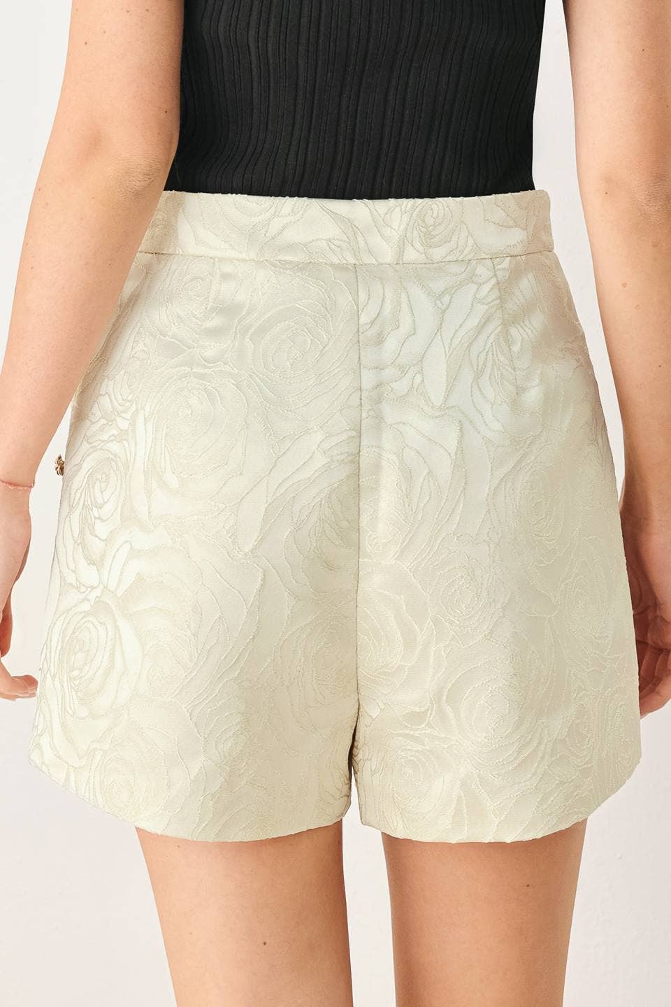 A close-up of a model wearing high-waisted, light beige textured shorts with a subtle embossed floral pattern, paired with a black ribbed top.