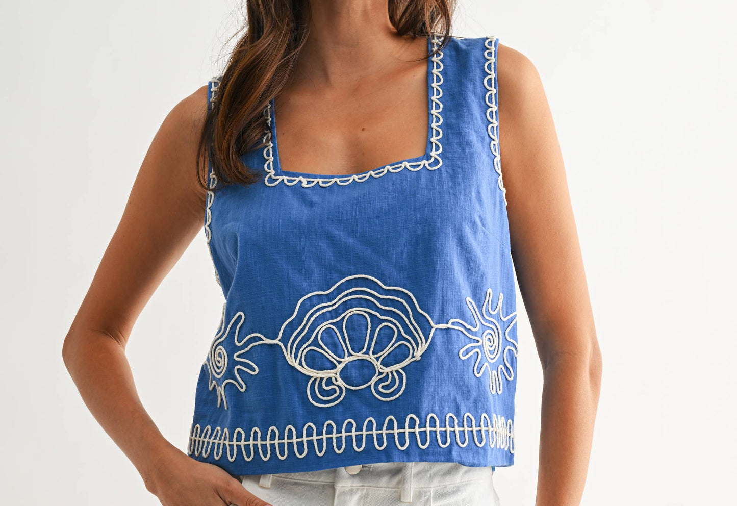 Woman wearing a blue tank top with white embroidery on a plain background