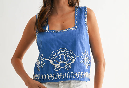 Woman wearing a blue tank top with white embroidery on a plain background
