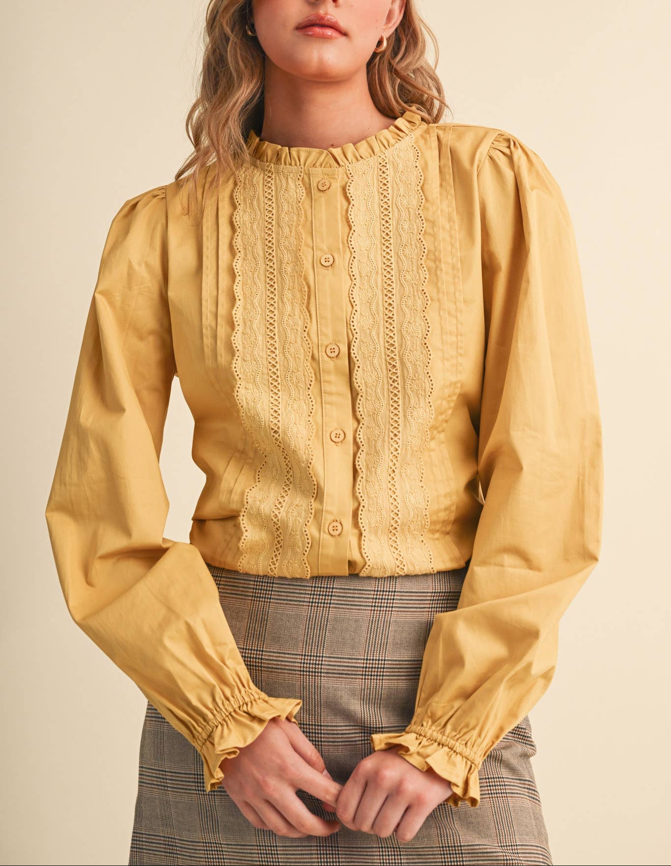 A woman wearing a mustard yellow long-sleeve blouse with a ruffled high neck, decorative vertical lace-like panels, and gathered cuffs, paired with a gray plaid skirt, against a plain beige background.