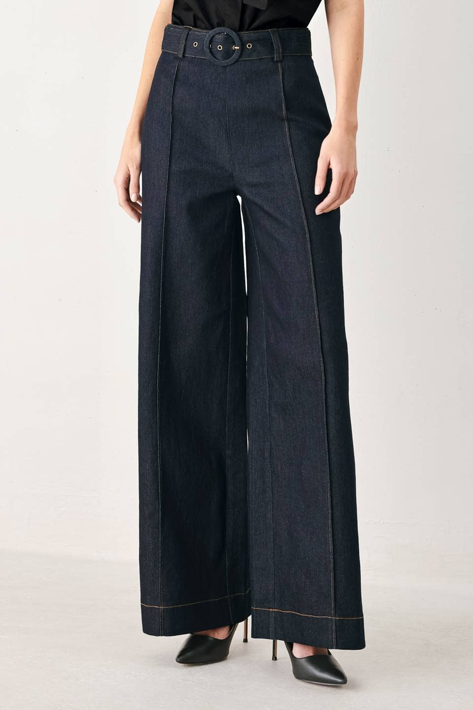 A model wearing dark indigo wide-leg denim pants with a matching belt, styled with black pointed-toe heels against a light gray studio background.