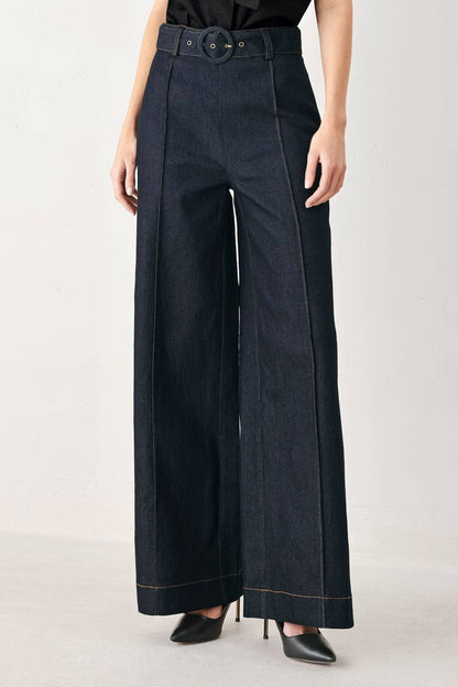 A model wearing dark indigo wide-leg denim pants with a matching belt, styled with black pointed-toe heels against a light gray studio background.