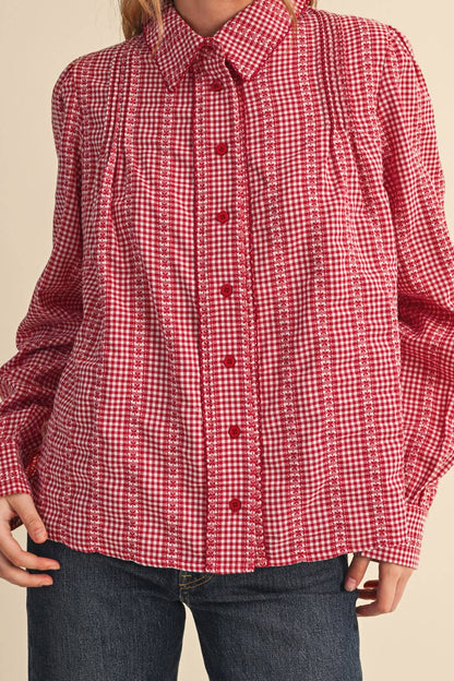A woman wearing a red and white gingham long-sleeve button-up shirt with pleated details on the chest and cuffs, paired with dark blue jeans, against a solid beige background.