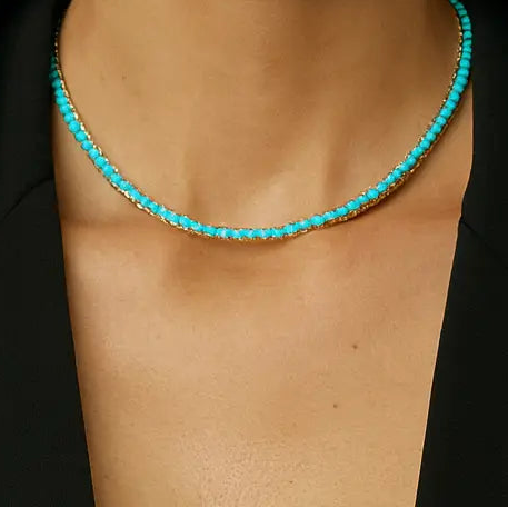 A close-up portrait of a woman wearing a delicate turquoise beaded necklace with a black blazer, set against a neutral textured gray background.