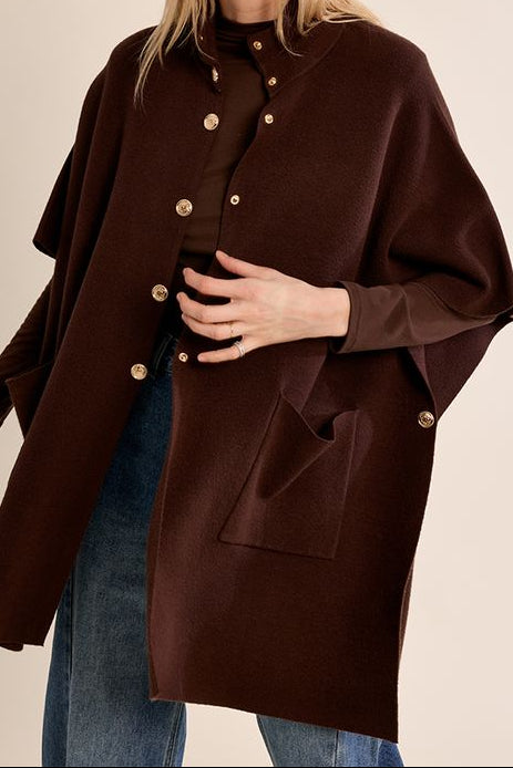 A woman modeling a dark brown cape-style coat with gold-tone buttons, paired with blue jeans, against a solid beige studio background.