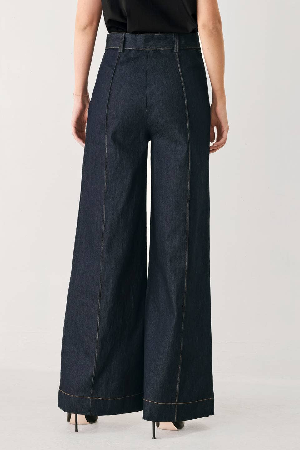 A model shown from the back wearing dark indigo wide-leg denim pants with contrast light stitching, paired with a black top and black heels, against a light gray studio background.