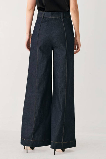 A model shown from the back wearing dark indigo wide-leg denim pants with contrast light stitching, paired with a black top and black heels, against a light gray studio background.