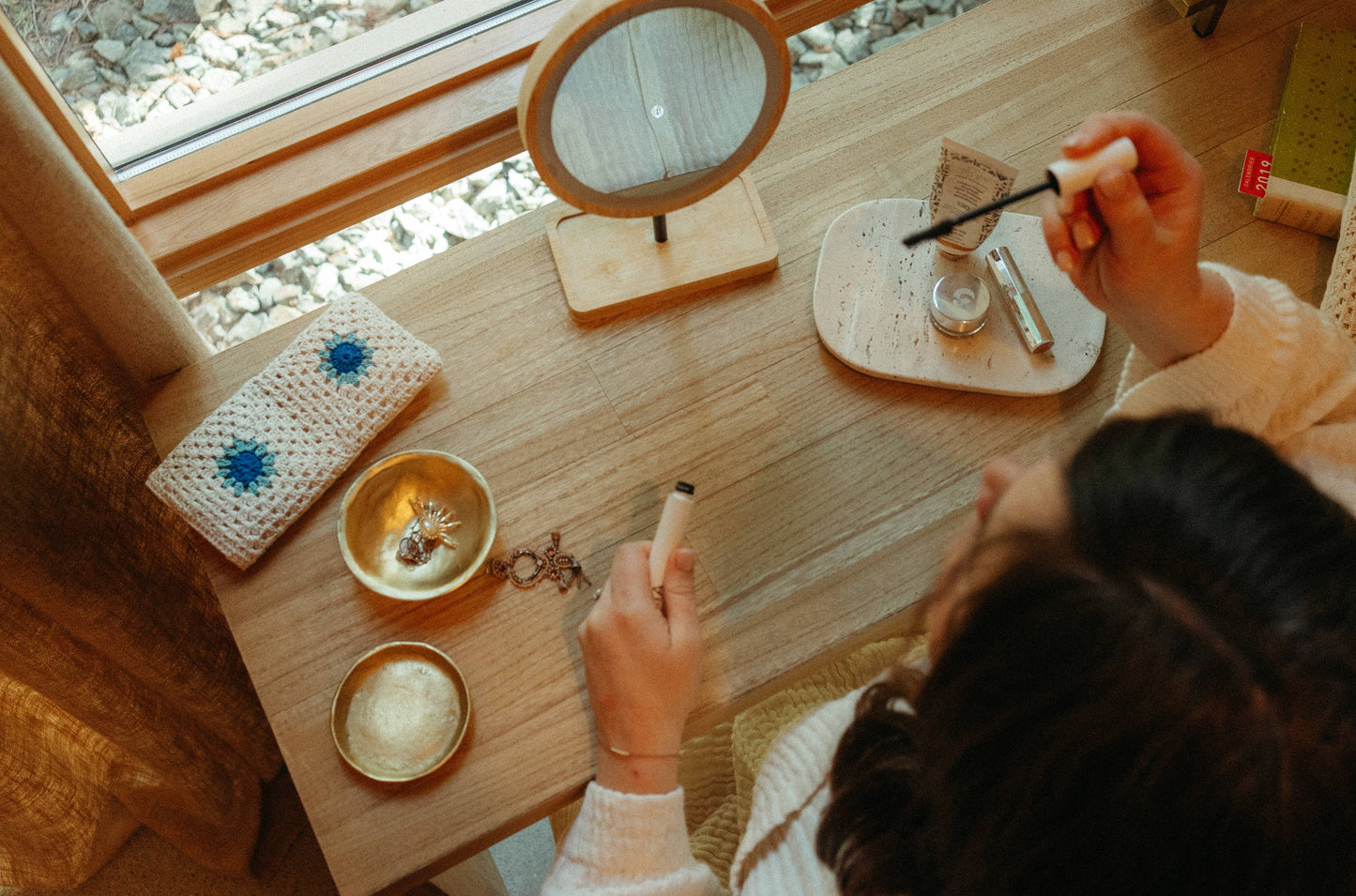 Person applying makeup at a wooden desk with various items including a mirror and small bowls.