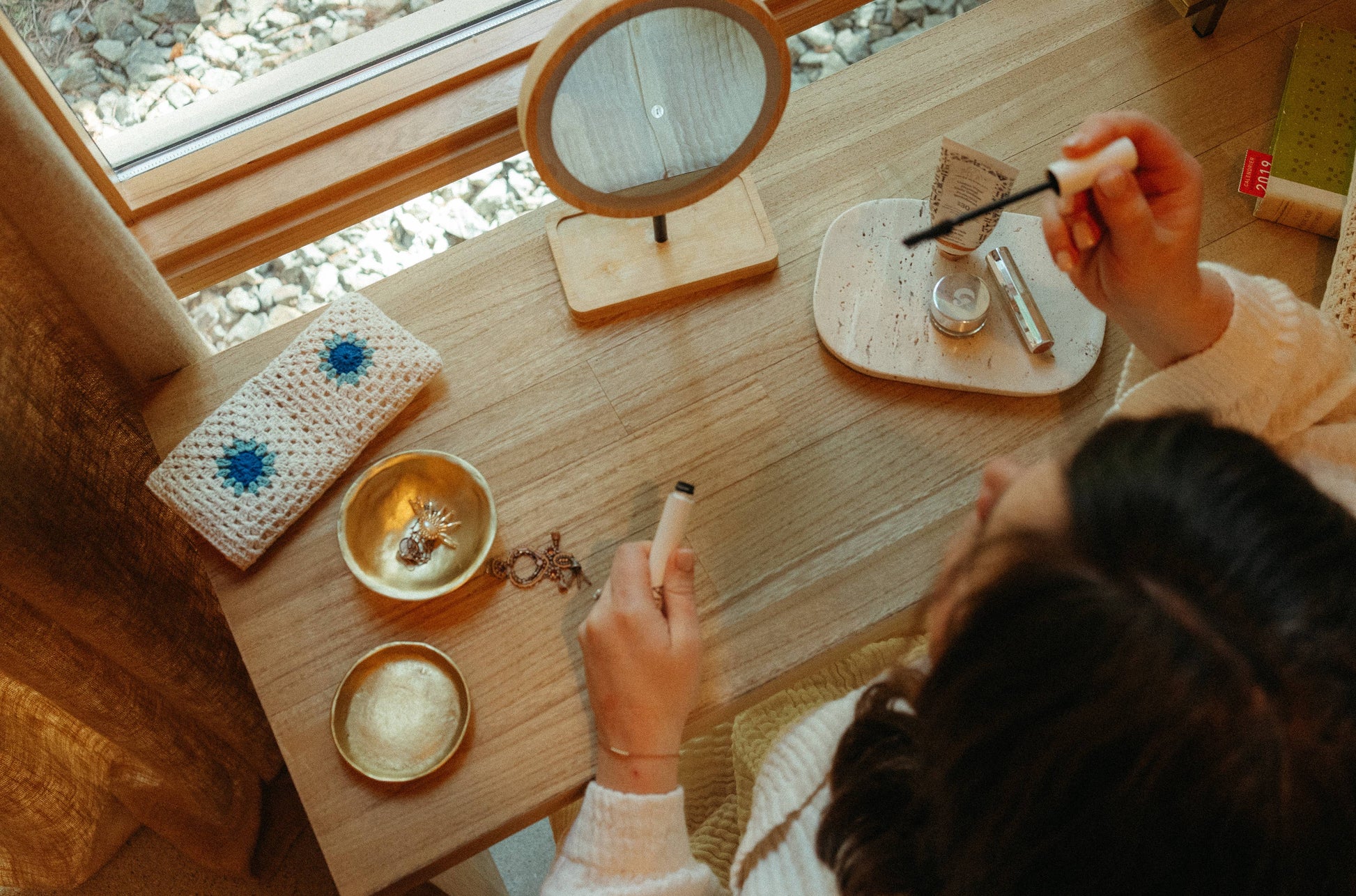 Person applying makeup at a wooden desk with various items including a mirror and small bowls.