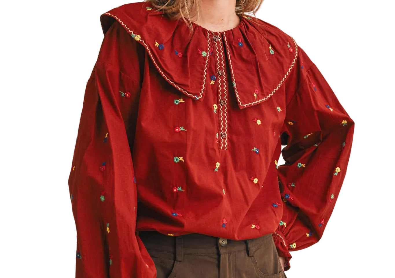 A woman wearing a red long-sleeve blouse with a large ruffled collar, embroidered details, and brown trousers, photographed against a plain white  studio background.
