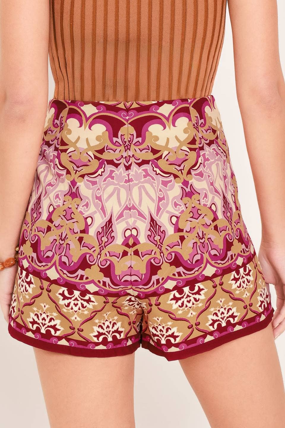A close-up of a model wearing high-waisted shorts with a detailed baroque-style damask pattern in shades of magenta, beige, and gold, paired with a brown ribbed top.