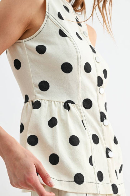 A close-up of a model wearing a sleeveless cream-colored shirt with large black polka dots, featuring a buttoned front and a gathered waist.