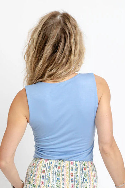 A woman shown from behind wearing a light blue sleeveless top and a long, tiered maxi skirt with a multicolor vertical stripe and floral pattern. She is standing against a plain white studio background.