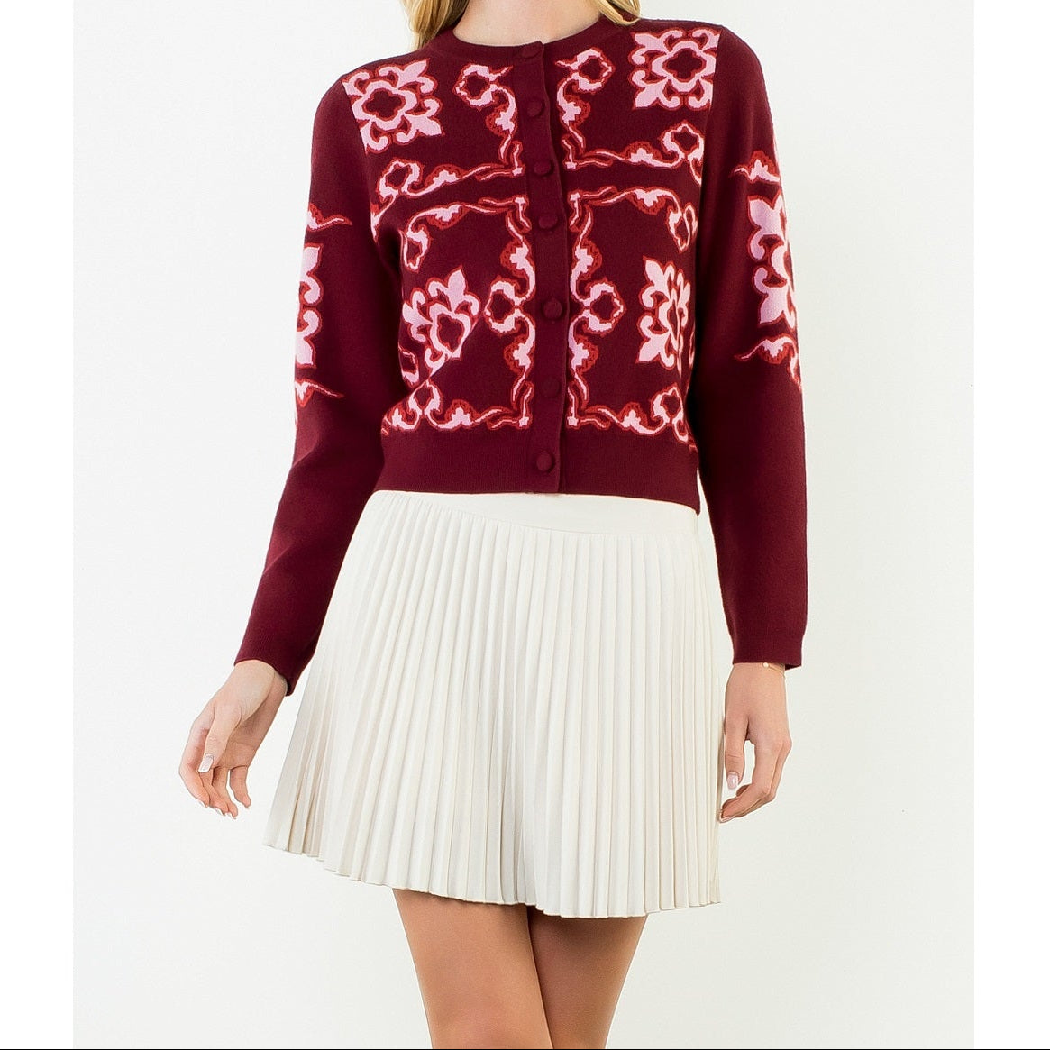 A woman modeling a burgundy cardigan with pink floral embroidery and a white pleated skirt, set against a plain white studio background.