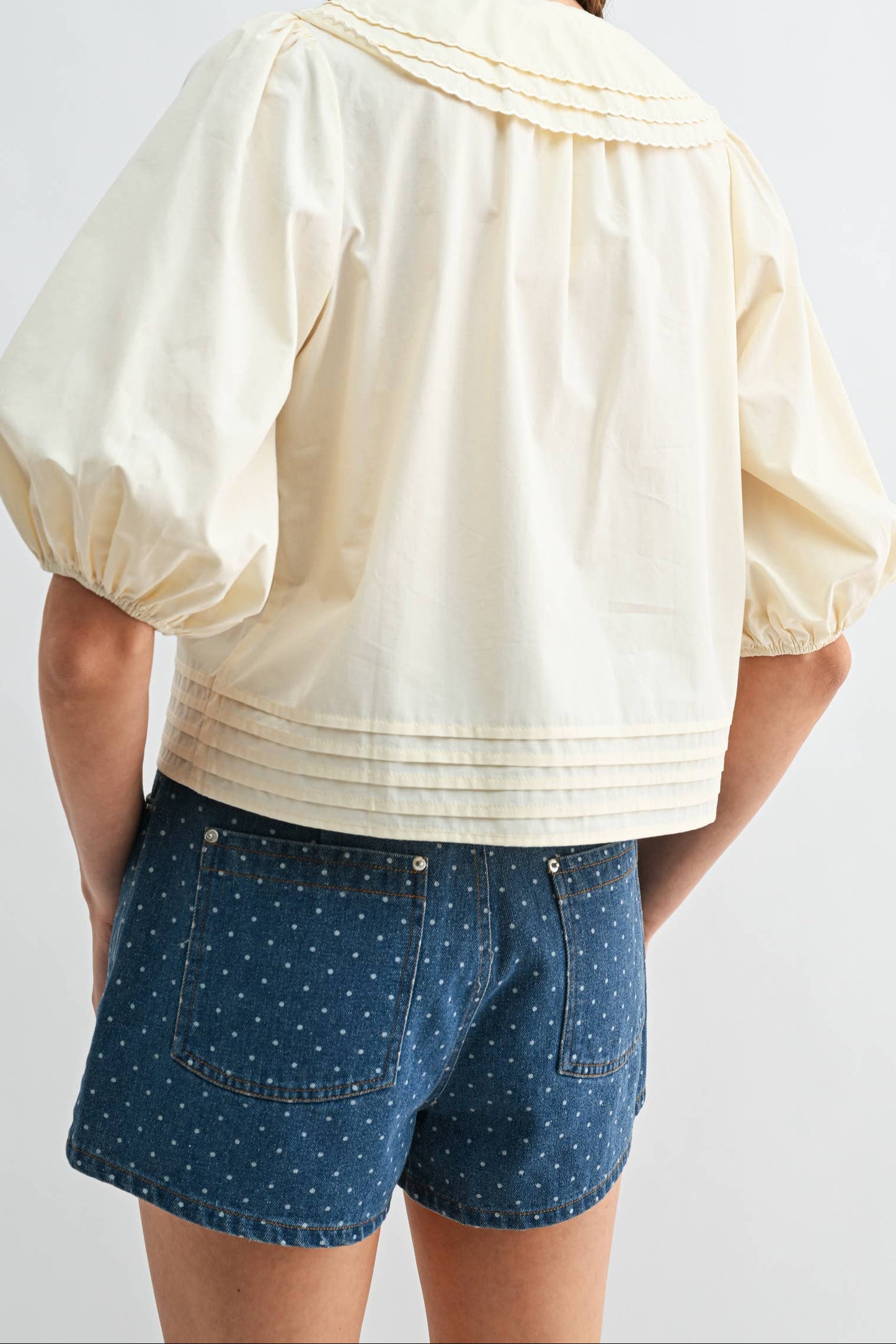 A woman shown from the back wearing a cream puff-sleeve blouse with pleated detailing and blue polka-dot shorts against a light gray studio background.