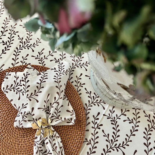 Fabric with brown leaf pattern on a white background, placed on a woven surface with blurred greenery in the background.