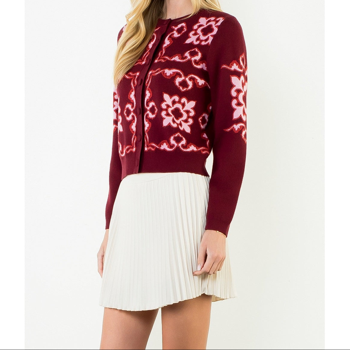 A woman modeling a burgundy knit cardigan with pink and white floral patterns, paired with a cream pleated mini skirt, against a plain white studio background.