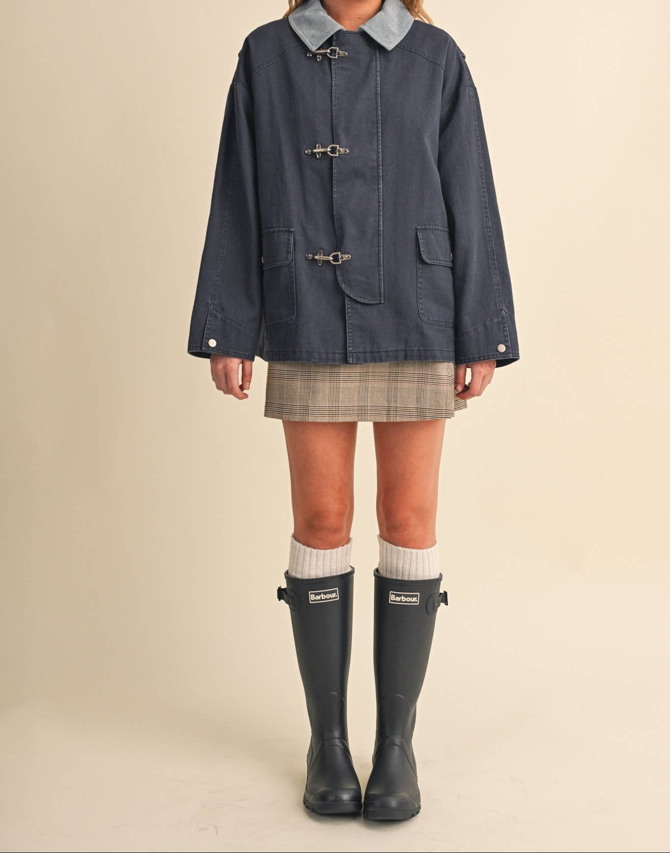 A model stands against a plain beige studio background wearing a dark navy blue coat with a light blue collar, a plaid mini skirt, white socks, and dark rain boots. The boots feature a visible 'HUNTER' brand label.