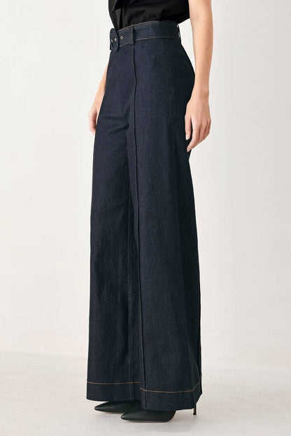 A model wearing dark navy blue wide-leg denim pants with a matching belt, styled with a black top and black pointed-toe heels against a light gray studio background.