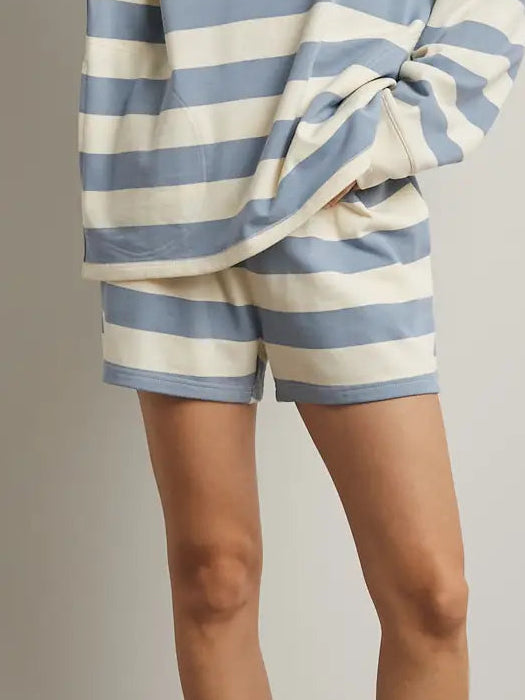 A woman modeling a matching two-piece loungewear set featuring a blue and off-white striped long-sleeve top and shorts, paired with white slide sandals, against a neutral studio backdrop.
