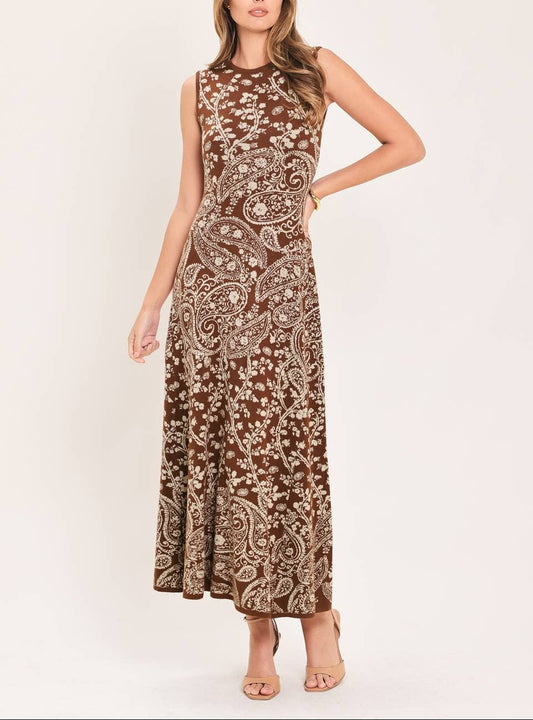 A woman modeling a sleeveless, high-neck maxi dress with a brown and beige paisley pattern, standing against a plain light beige studio background.