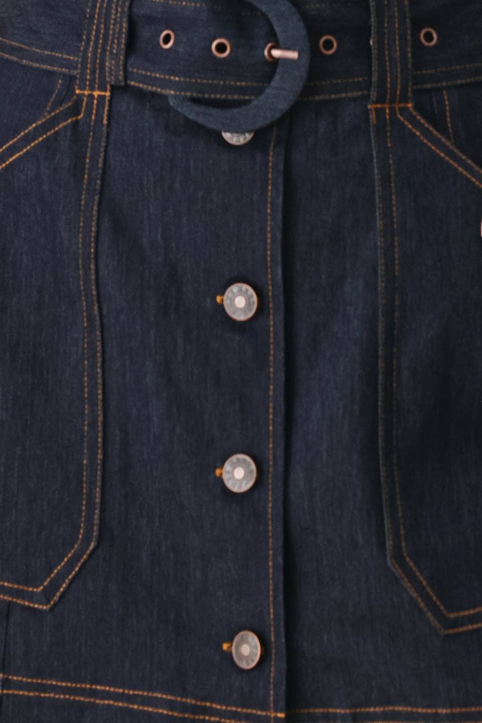 Close-up of a dark indigo denim garment showing the front button closure, belt loops, and contrast orange-yellow stitching.