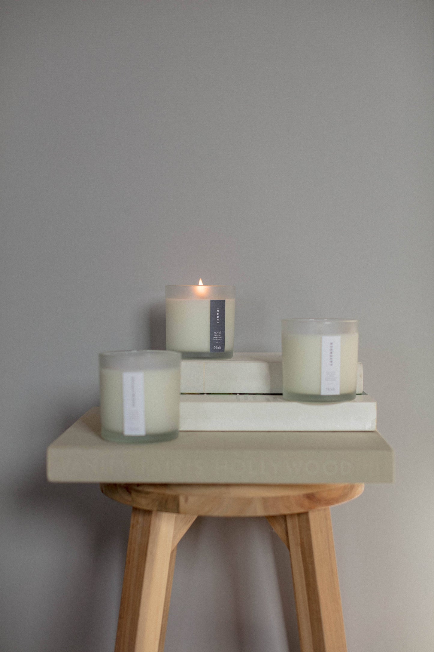 Three candles on books with a small candle lit on a wooden stool against a gray wall.
