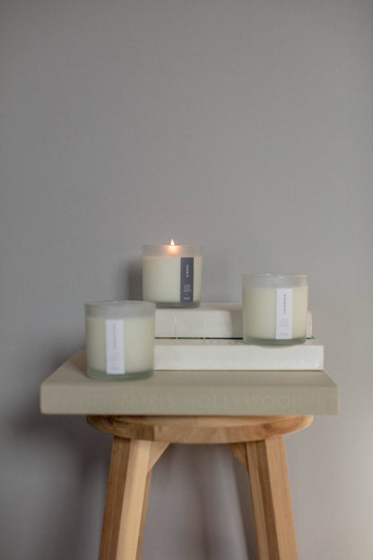 Three candles on books with a small candle lit on a wooden stool against a gray wall.