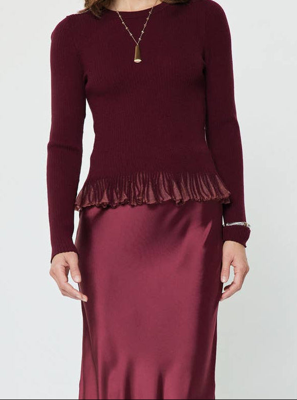 A woman modeling a long-sleeve burgundy knit top with a fringed hem and a matching satin midi skirt, paired with dark brown ankle boots, against a plain light gray background.