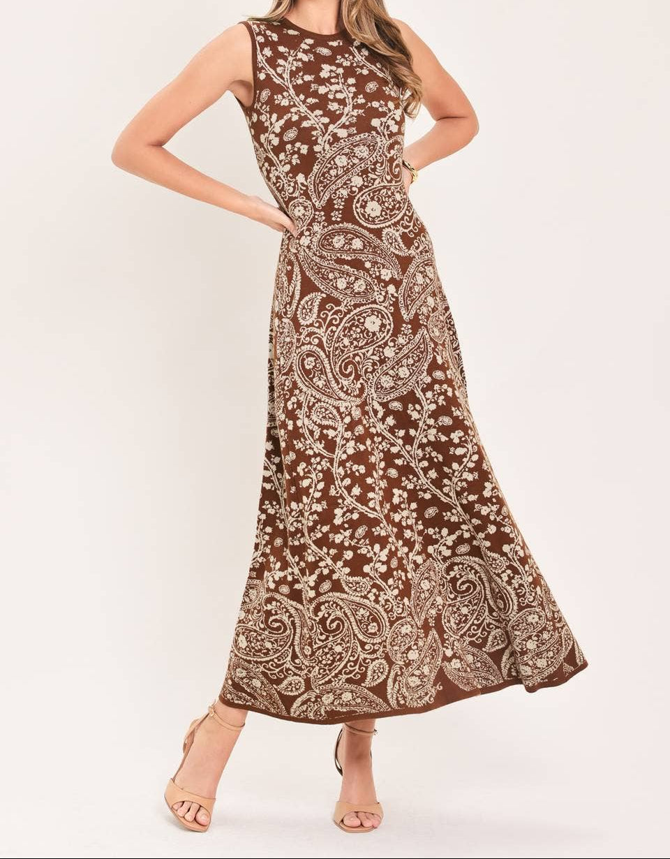 A woman modeling a sleeveless, floor-length brown maxi dress with an intricate beige paisley and floral pattern, posed against a solid off-white studio background.