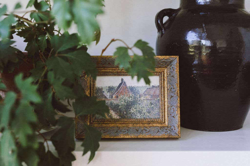 Small framed painting on a shelf with a dark vase and green leaves in the foreground
