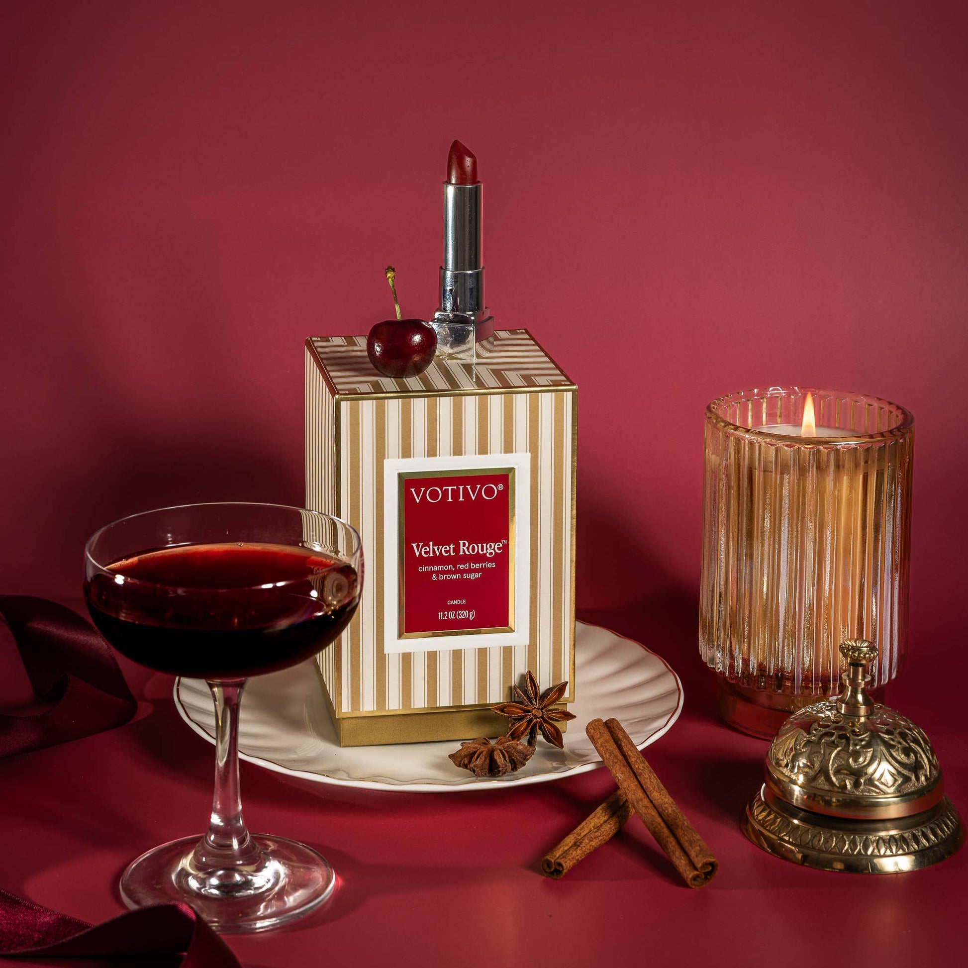 Votivo Velvet Rouge perfume box displayed on a white plate with a lipstick, cherry, glass of red wine, cinnamon sticks, star anise, and a lit candle against a deep red background.