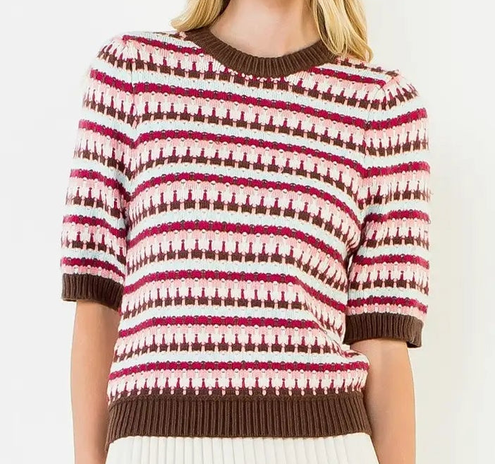 A woman modeling a short-sleeve knit sweater with horizontal brown, white, and pink geometric patterns, paired with a white pleated mini skirt, against a plain white background.