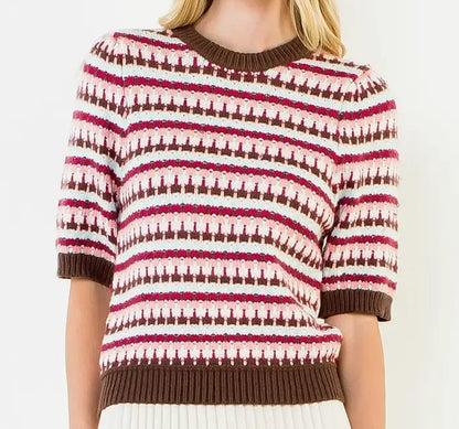 A woman modeling a short-sleeve knit sweater with horizontal brown, white, and pink geometric patterns, paired with a white pleated mini skirt, against a plain white background.