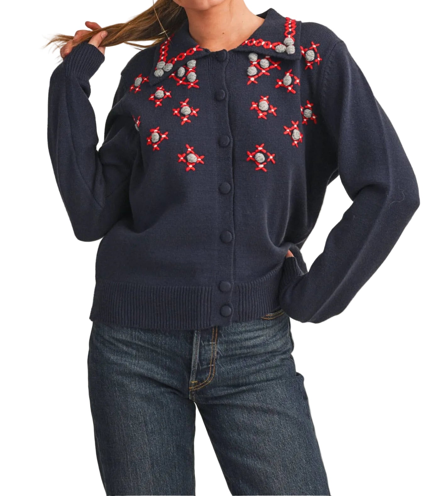 A woman modeling a navy blue knit cardigan with red and white embroidered geometric patterns across the chest and shoulders, paired with blue jeans against a solid beige background.