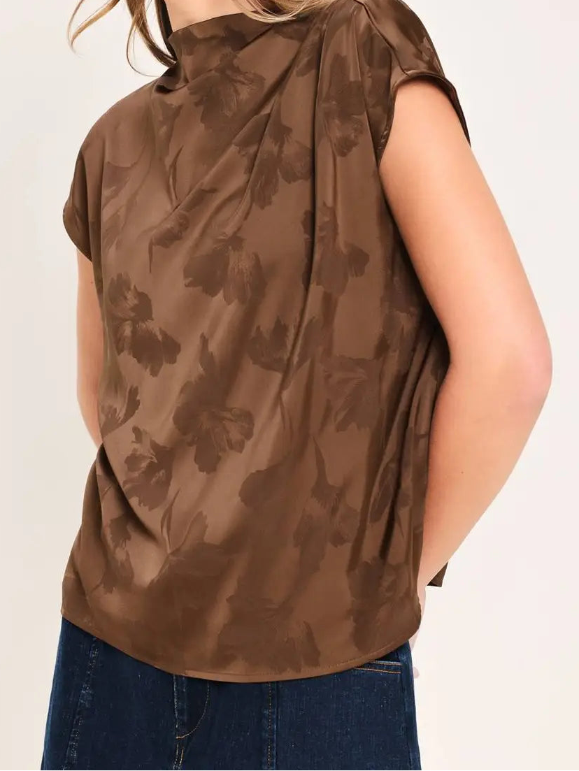 A woman wearing a brown short-sleeve top with a tonal floral pattern, paired with blue jeans, shown from a side profile against a light beige background.