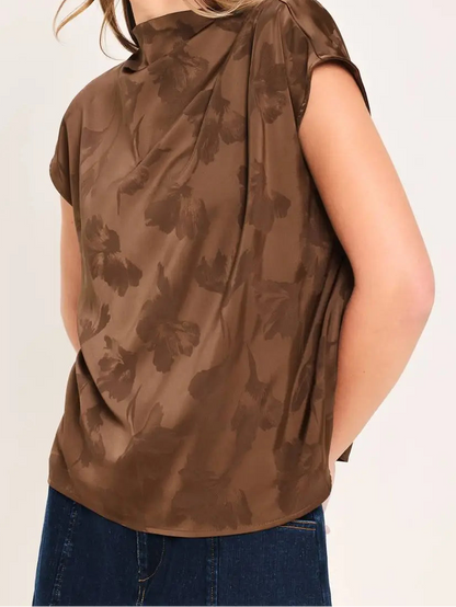 A woman wearing a brown short-sleeve top with a tonal floral pattern, paired with blue jeans, shown from a side profile against a light beige background.