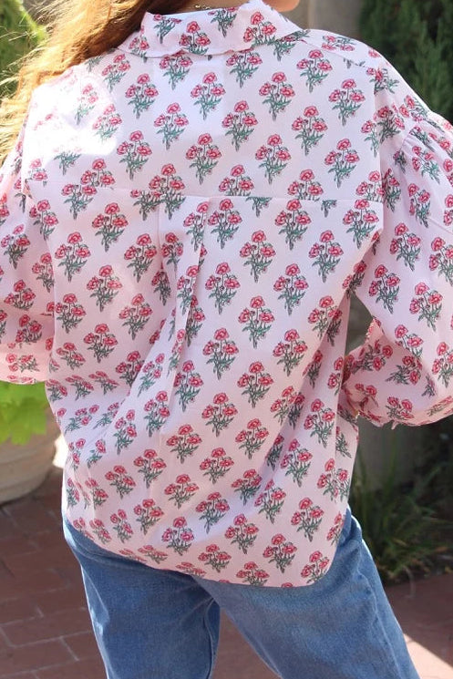 A woman viewed from behind wearing a light pink button-up shirt with a red and green floral block print, paired with blue jeans, standing outdoors in front of green plants.
