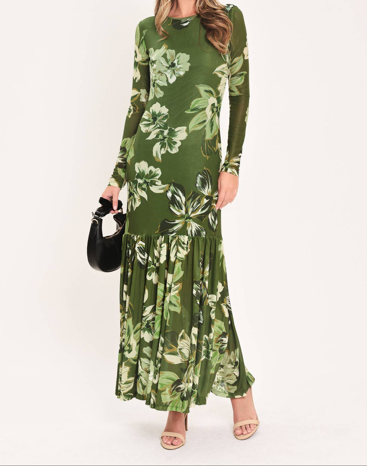 A woman modeling a long-sleeve olive green maxi dress with a large white and light green floral print, holding a small black handbag against a plain white studio background.