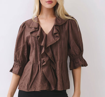 A blonde woman wearing a brown ruffled blouse with puff sleeves and black pants, standing against a light gray studio background.