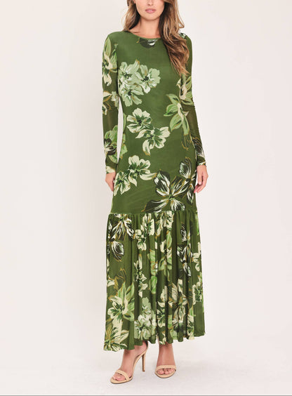 A woman modeling a long-sleeve maxi dress with a green and white tropical floral print, standing against a plain light beige studio background.