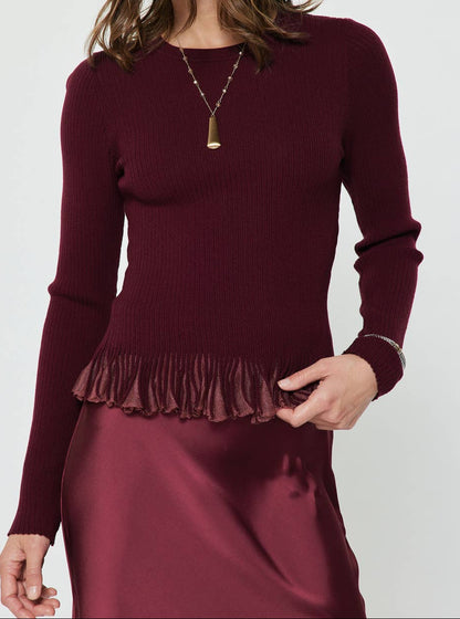 A woman wearing a ribbed long-sleeve knit sweater in deep burgundy with a ruffled hem, paired with a matching satin skirt, against a light gray studio background.