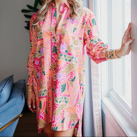 Woman wearing a colorful floral dress standing by a window indoors.