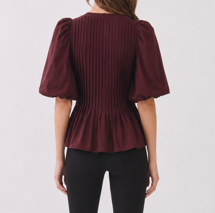 A woman viewed from the back wearing a burgundy pleated short-sleeve peplum top and black cropped pants with black mule heels, against a light gray studio background.