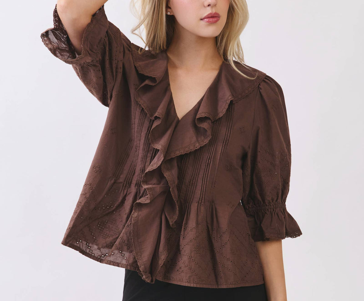 A woman modeling a brown ruffled blouse with puff sleeves and black pants against a light gray studio background.