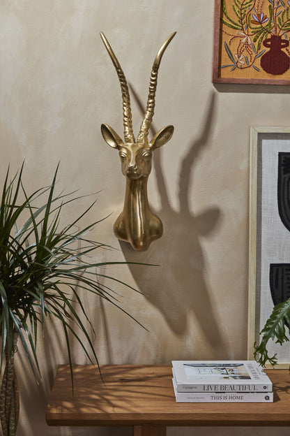 Gold antelope head wall decoration on a beige wall with books and a plant on a wooden surface.