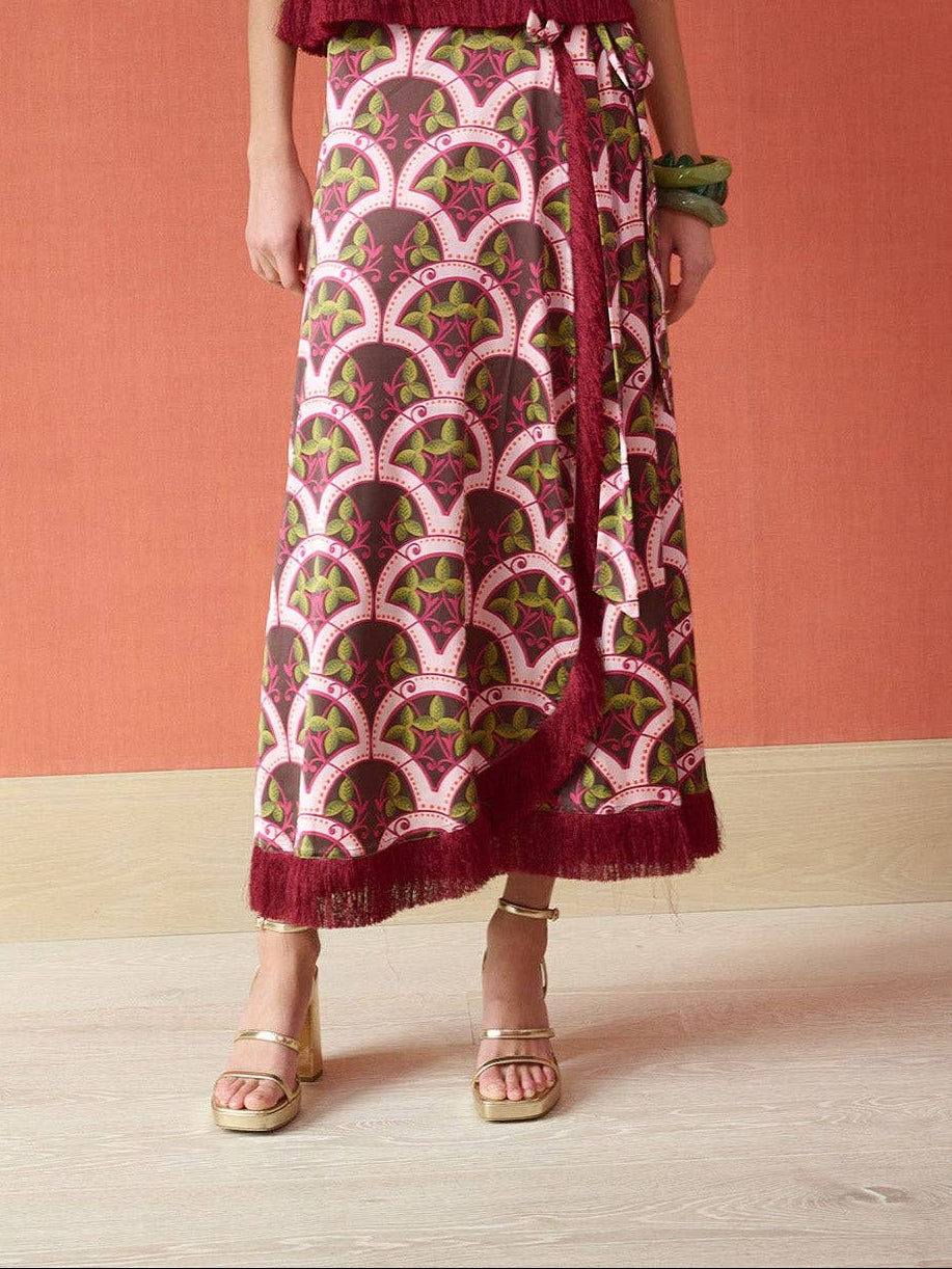 A woman stands against a terracotta-colored wall wearing a long pink and burgundy patterned dress with a matching belt and gold sandals.