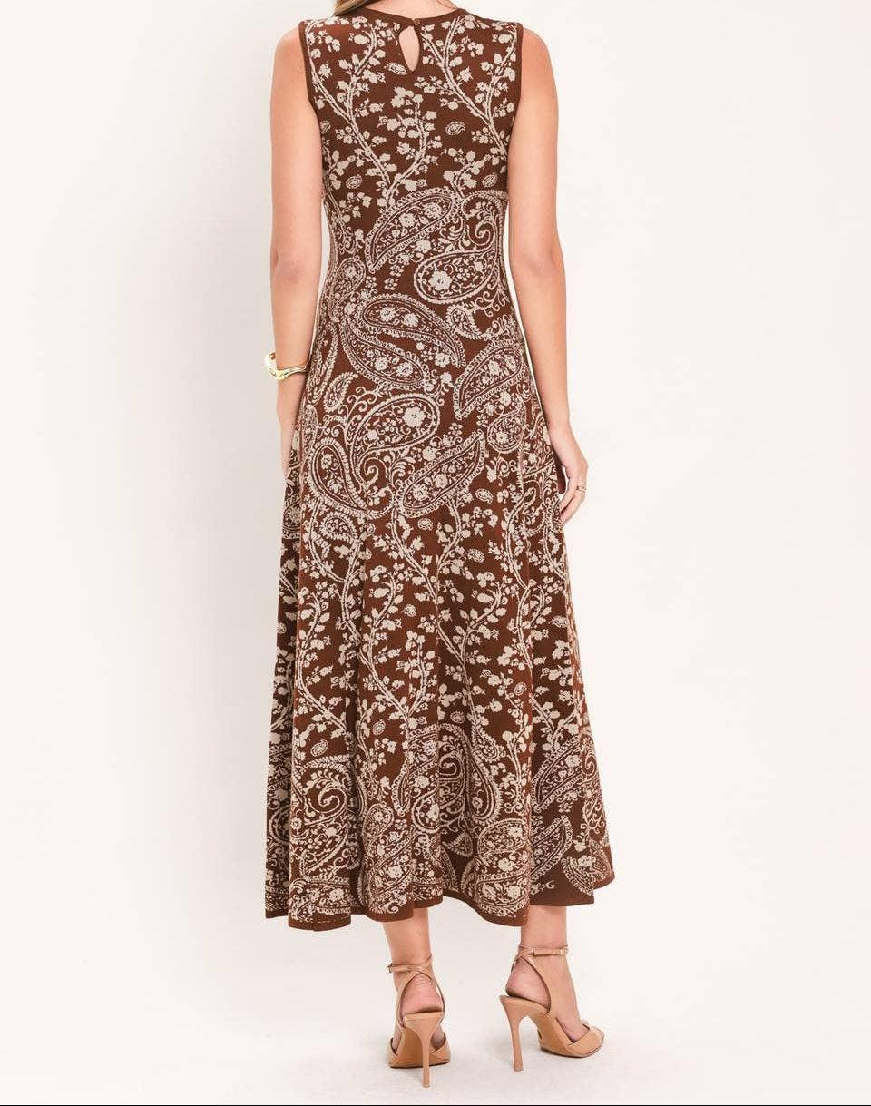 A woman viewed from the back wearing a sleeveless brown maxi dress with a white paisley pattern and a small keyhole cutout at the back, paired with beige high-heeled sandals against a plain white background.