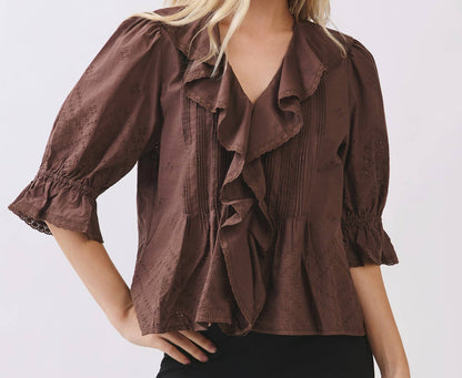 A blonde model wearing a brown ruffled blouse with puff sleeves and black pants, photographed against a light gray studio background.